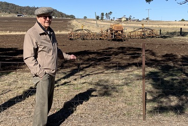 An elderly gentleman stands in front of a paddock, a seeder machine can be seen in the background